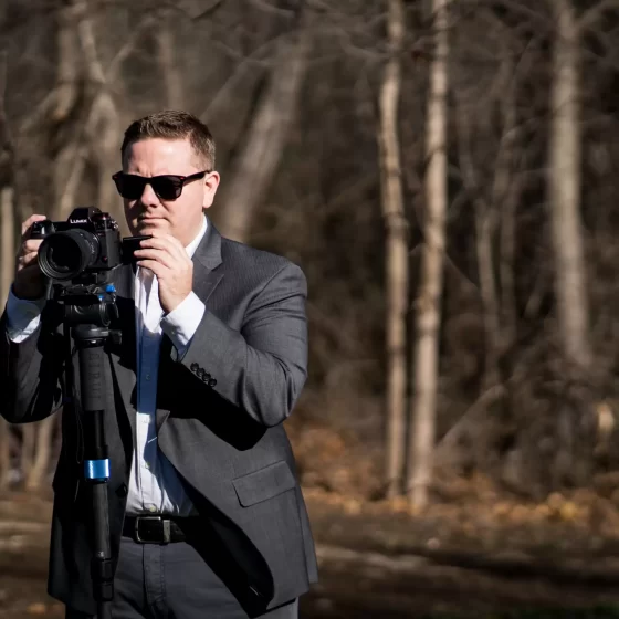 young man wearing sunglasses and suit jacket looks at display screen on camera on monopod as he stands in a wooded area