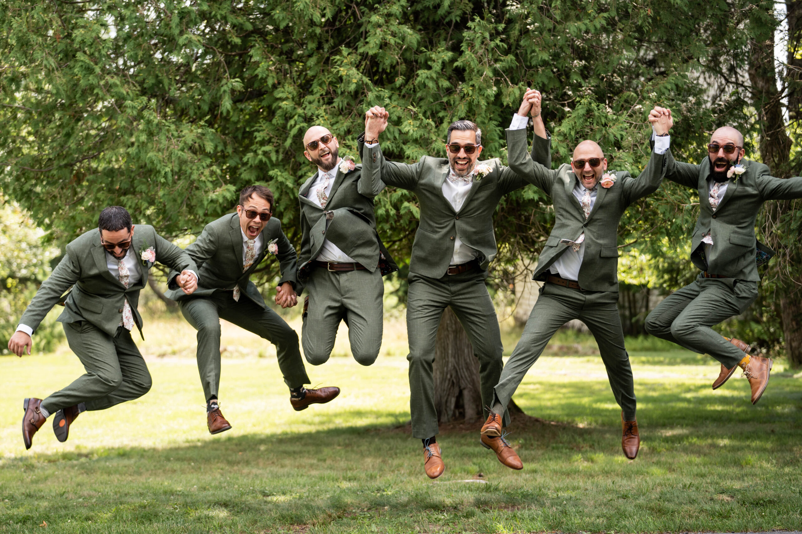groom and five groomsmen dressed in forest green suits and pink boutonnieres jumping in midair while smiling and laughing at the camera with green trees and grass in the background