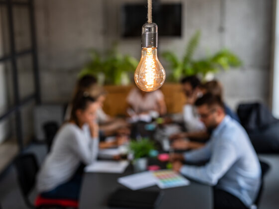 a lit lightbulb hanging from the ceiling in focus with a table of seven men and women seated at a conference table littered with documents as the out of focus background
