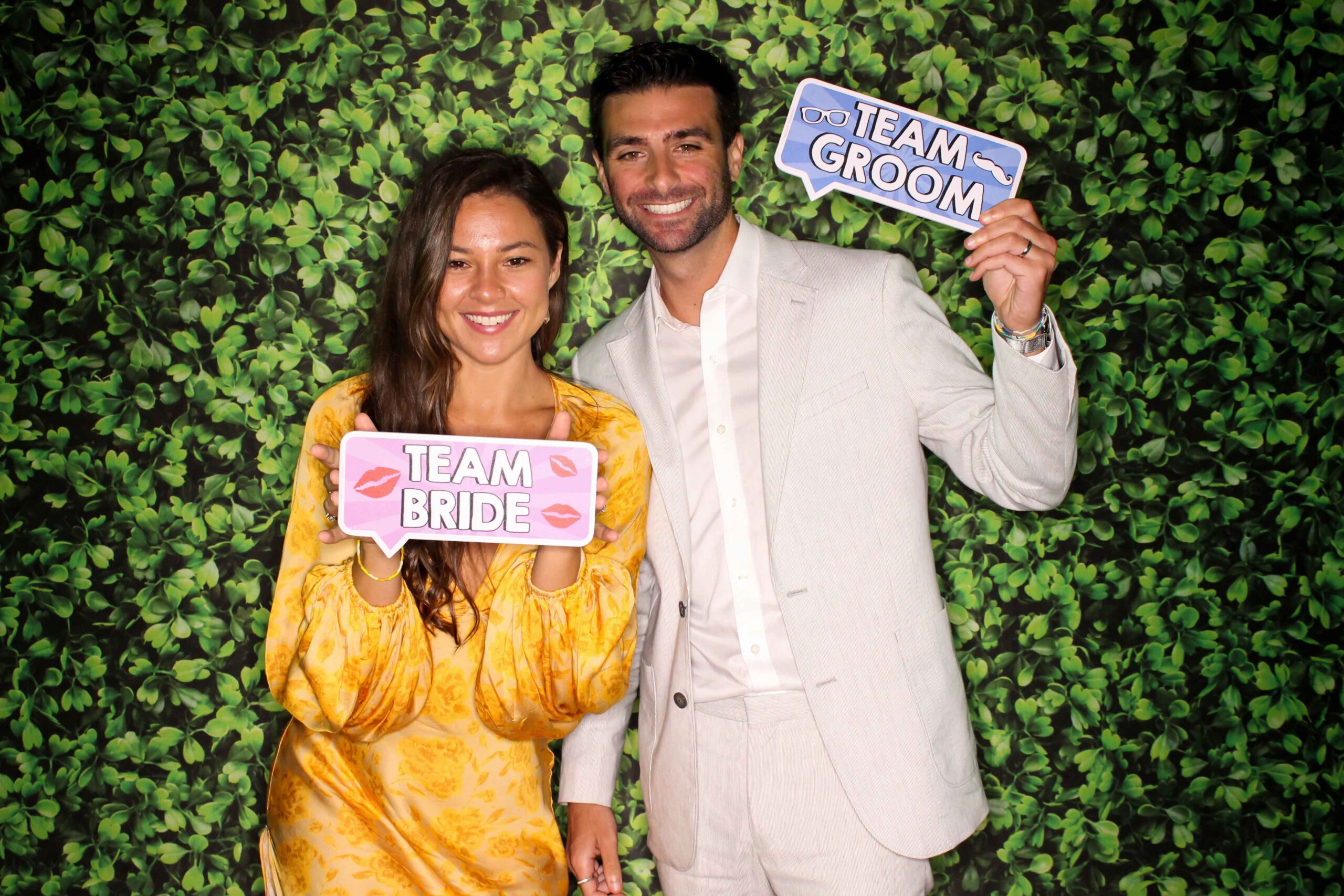 man holds a sign that says "team groom" and woman holds a sign that says "team bride" as they pose for a photo booth picture with a bright green hedge backdrop