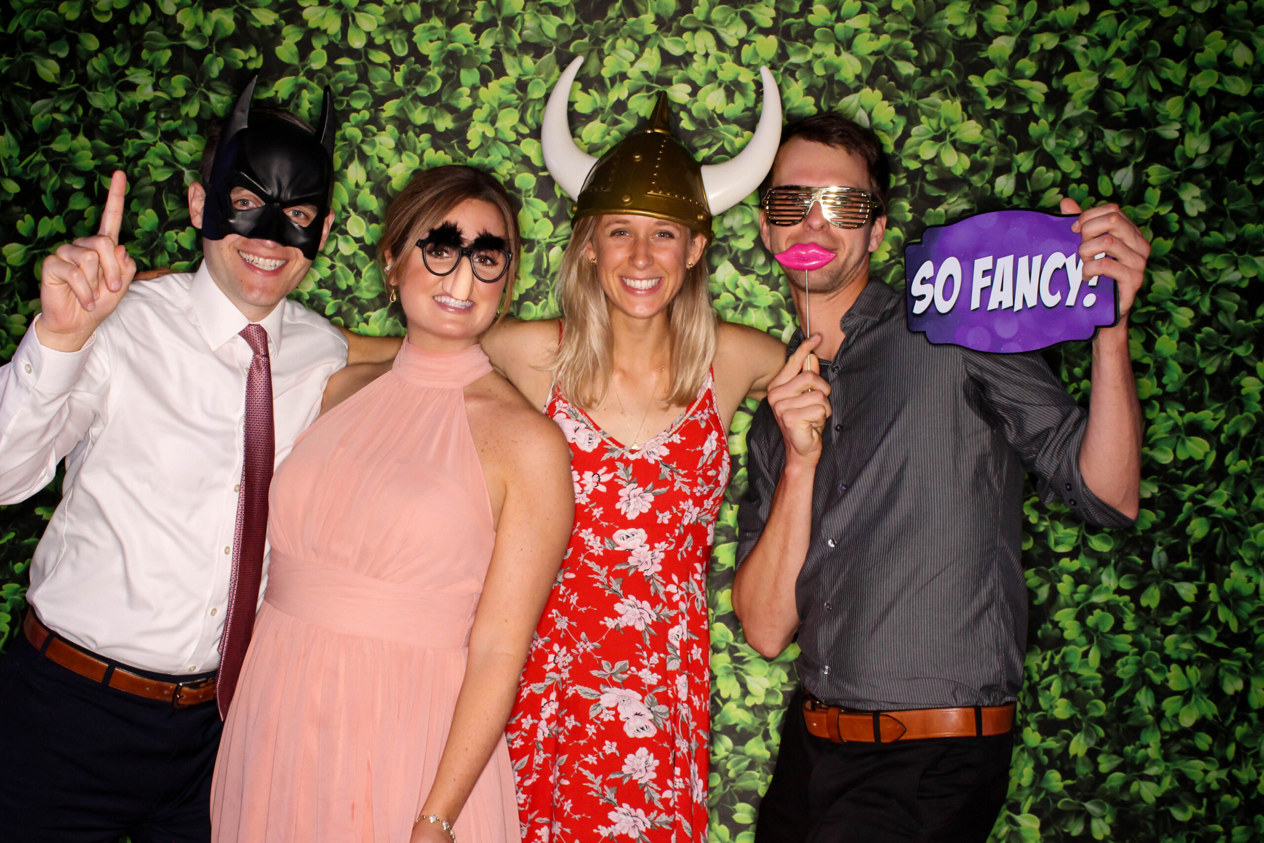 photo booth picture of four wedding guests in formal attire hugging on each other and wearing funny props against a green hedge background