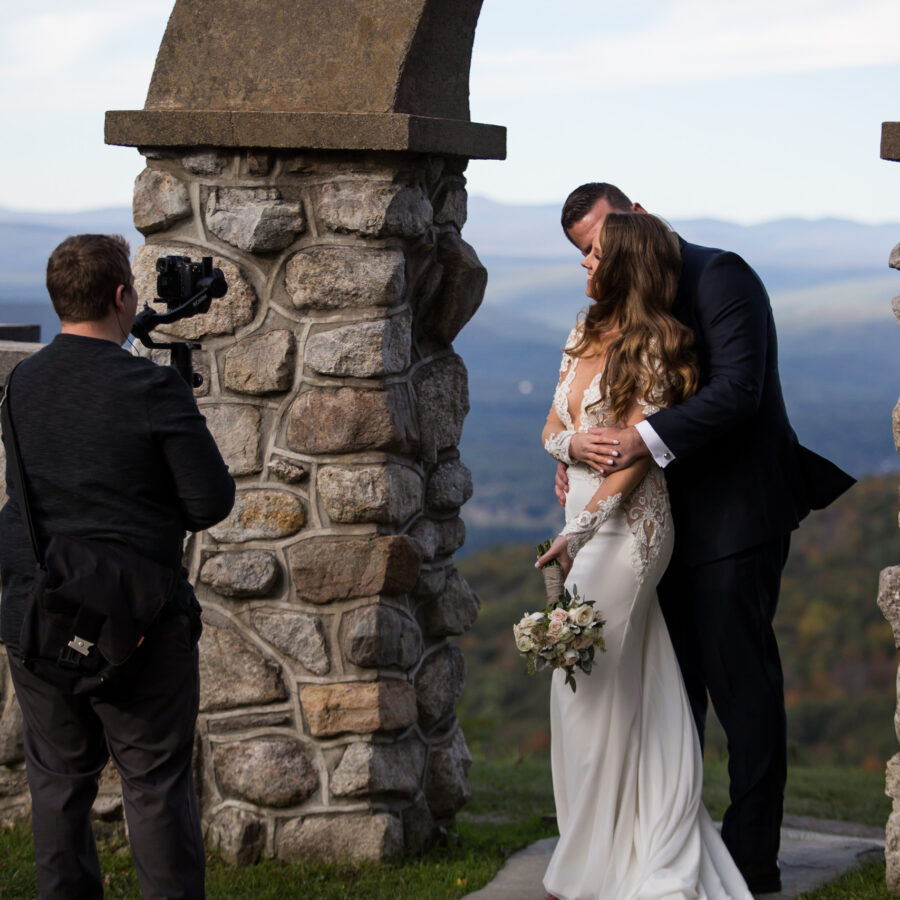 man getting video footage on gimbal as bride and groom pose under a stone arch with skies and mountain ranges in background