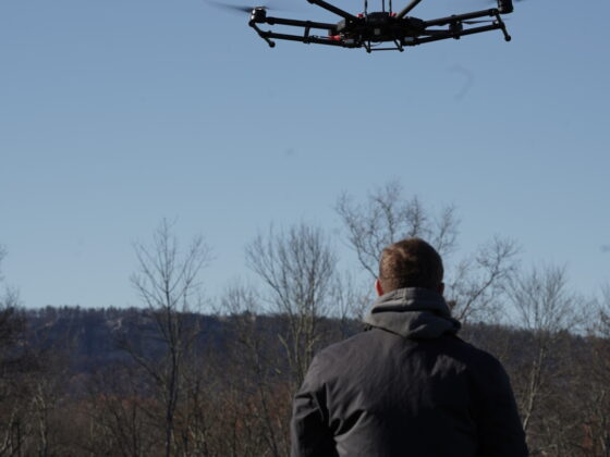 man facing away from the camera as he controls a large drone flying above him in clear blue skies with trees in the background