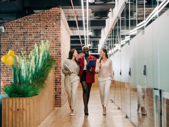 three business women smile and talk to each other as they walk through the hallway of their workplace which looks modern with large glass doors and exposed brick walls