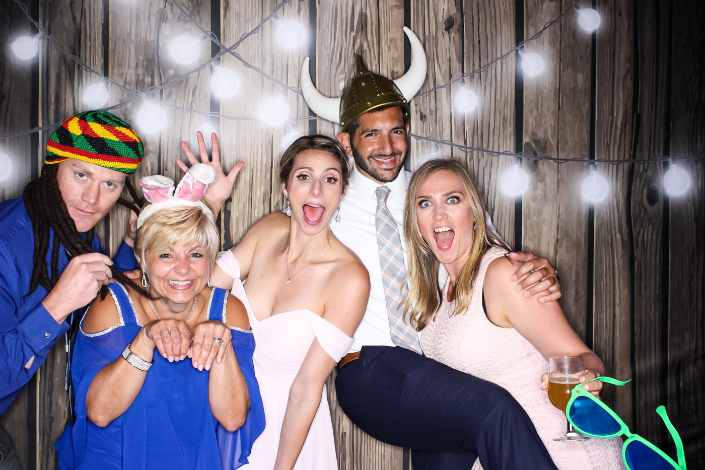 photo booth picture of a bride and groom with their family and friends holding each other and making funny faces while wearing silly props against a wood backdrop with string lights
