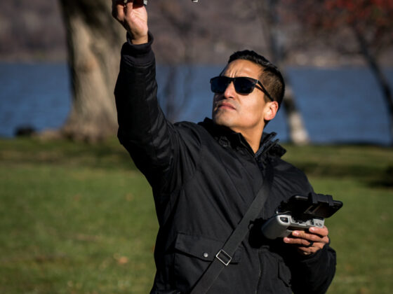man wearing sunglasses has a drone remote in one hand while looking up at a descending drone that he is catching in his other hand