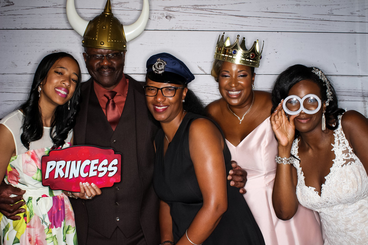 photo booth picture of bride and four of her friends and family wearing silly props and holding up funny signs against a white washed wood background