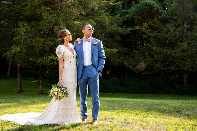 bride resting her arm on groom's shoulder while both wear sunglasses and stare into the left of a clearing in a forest