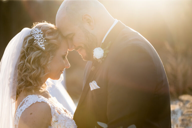 bride and groom standing forehead to forehead as both smile and close their eyes with a sun flare in the background