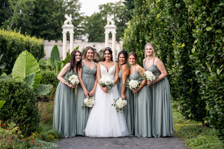 bride in wedding gown holding a bouquet surrounded by 5 bridesmaids holding their bouquets and smiling at a camera while everyone stands in lush green gardens with ornate white stone pillars in the background