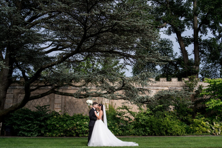 wide angle shot with bride and groom kissing in the middle of lush green gardens with enormous trees and an ornate stone wall in the background