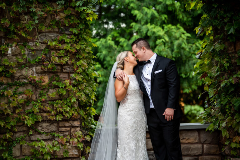 bride and groom stand in front of an ivy covered stone wall while smiling with eyes closed and noses touching