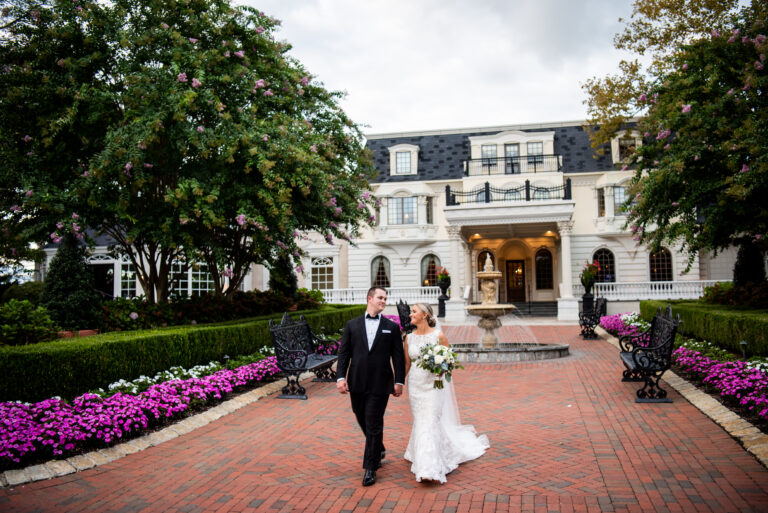 smiling bride and groom walking away from a large white mansion along a brick pathway lined with bright pink and white flowers and manicured grounds