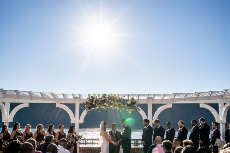 wide shot of bride and groom facing each other and holding hands during their ceremony at The Grandview in Poughkeepsie, NY with clear blue skies and the Hudson River in the background