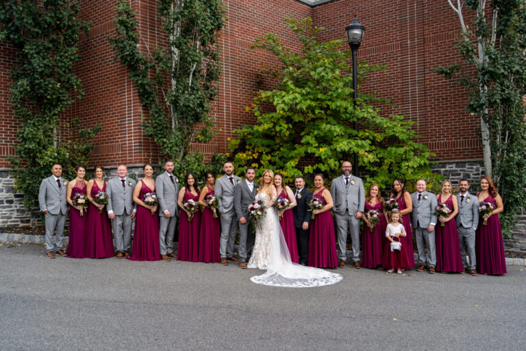 bride and groom standing in the middle as bridal party of twenty stands on either side with a brick building and trees in the background