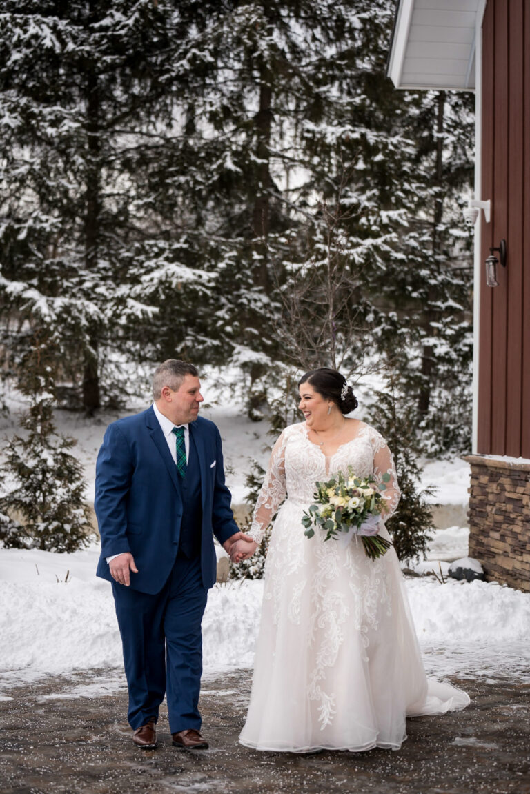 bride and groom standing outside in front of snow covered pine trees and snowy sidewalks holding hands while looking at each other and smiling