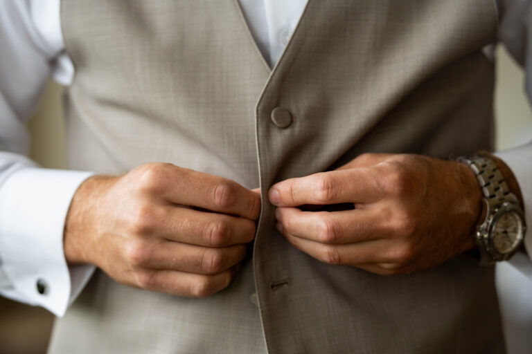 close up of groom's hands buttoning the second button of his high quality vest while sporting a luxury watch on his left wrist