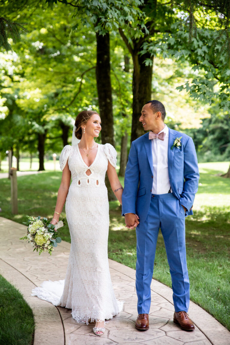 Bride and groom holding hands while looking and smiling at each other and walking on a stone path in a park with trees and sunlight