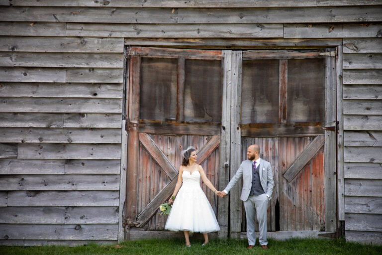 bride wearing a vintage tulle midi dress and vintage face veil smiling and holding groom's hand in front of a rustic barn door