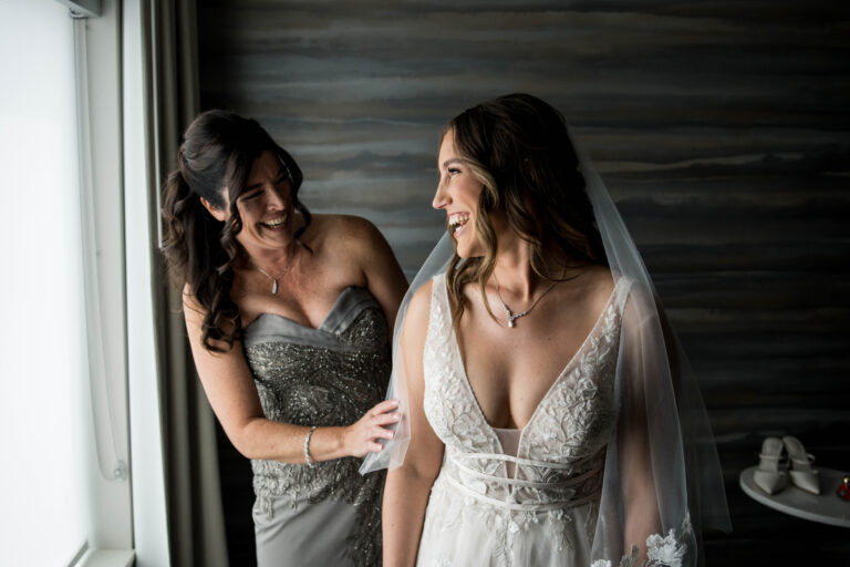 bride and mother standing by the window and looking at each other while laughing as mother adjusts veil