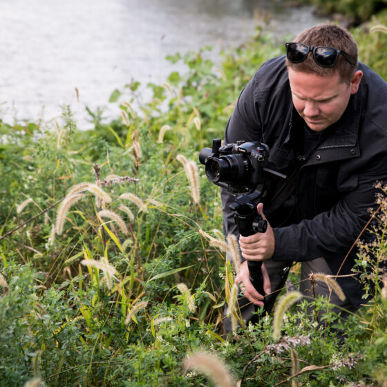 videographer crouching in tall grass near a body of water while filming with a camera on a monopod