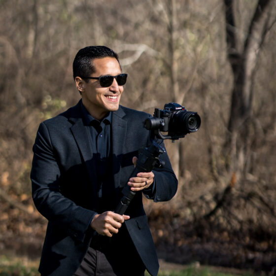 videographer holding a gimbal on a sunny day in a forest while wearing sun glasses and smiling