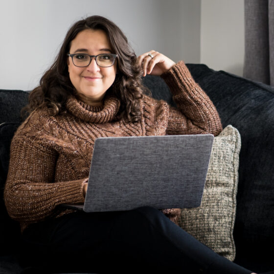 smiling woman wearing glasses and brown wool sweater seated on a large black couch with a silver laptop on her lap