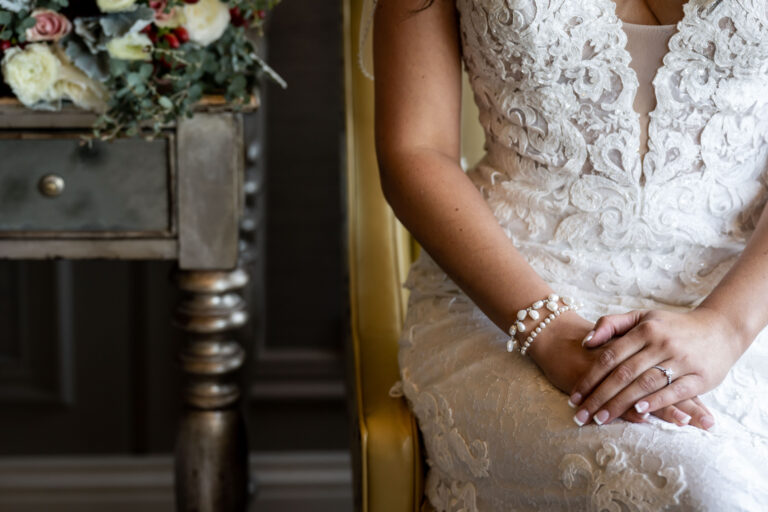closeup of bride's hands crossed on her lap while wearing her gown as she sits in an armchair next to a vintage side table on which her bouquet is placed