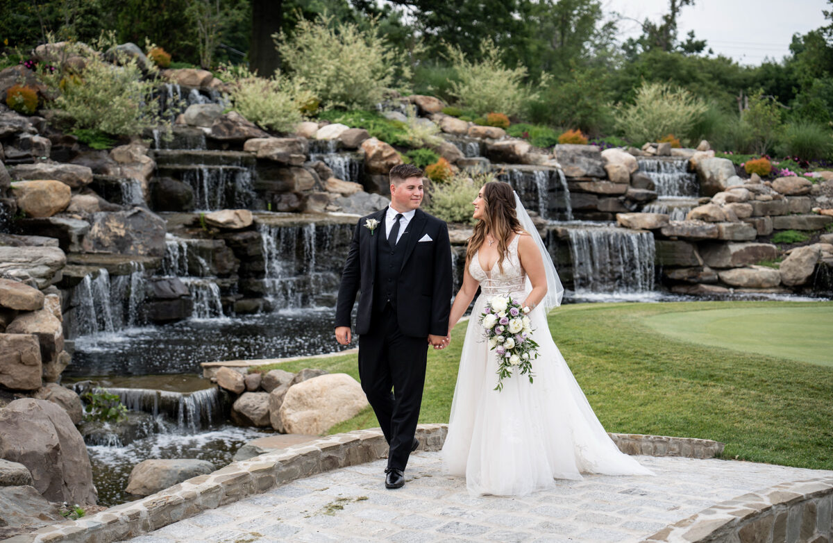 bride and groom hold hands and smile at each other as they walk over a stone bridge surrounded by manicured gardens and cascading waterfalls