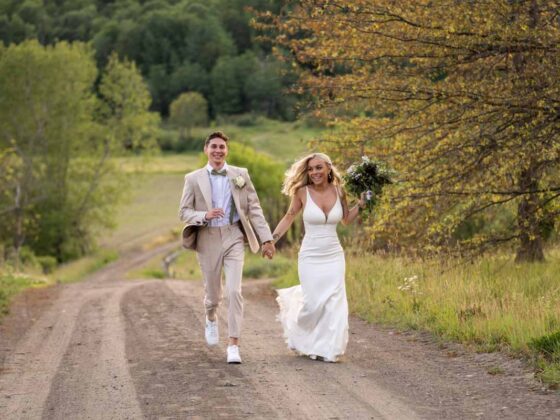 newly married bride and groom hold hands and run together along a dirt road lined with grass and trees; both are smiling widely and the bride is holding a bouquet; rays of light during golden hour shine through the bride's wavy blonde hair